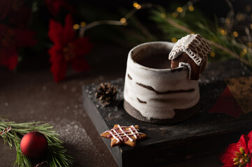 Hot winter drink in a clay mug. Close-up of hot chocolate with cinnamon stick, gingerbread house. Winter cozy still life with cocoa, Christmas decorations, fir branch and lights. © Lizaveta