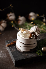 Hot winter drink in a clay mug. Close-up of hot chocolate with cinnamon stick, gingerbread house whipped cream. Winter cozy still life with cocoa, Christmas decorations, fir branch and lights. © Lizaveta
