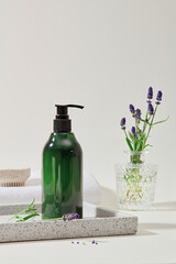 Close-up of a labelless transparent green shampoo bottle displayed on a gray tray, placed next to a vase of lavender flowers, standing out against a white background. Space for ads.