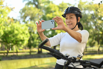 Happy middle age woman in bike helmet stops to taking photo of landscape with mobile phone.