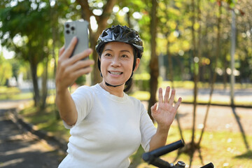 Cheerful middle age woman having video call on mobile phone while resting from cycling in a green...