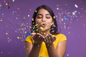 An indu girl blowing a bunch of confetti that she holds in her hands on a solid colored purple background. celebrating