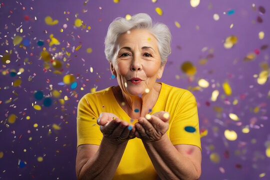 An Older Woman Blowing A Bunch Of Confetti That He Holds In His Hands On A Solid Colored Purple Background