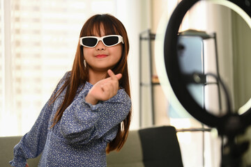 Cheerful Asian girl in sunglasses dancing in front of phone recording video for social network.