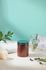 A brown jar of scrubs is displayed on the table with cotton towels, green leaves, a candle and lavender flowers. Skin care with products containing natural ingredients