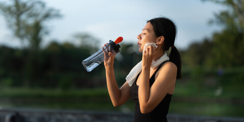 Beautiful young Asian woman stops to drinking water and wipe off her sweat after her evening run at...