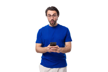 A 30 year old European guy with black hair and a beard in a blue T-shirt uses a smartphone and looks at the camera in surprise
