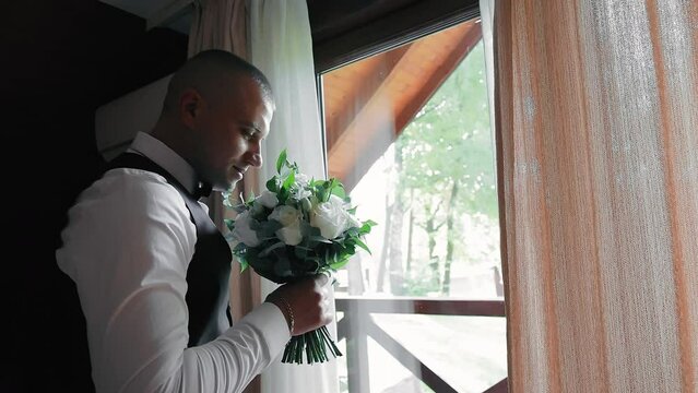 Grums Anticipation with Buchuet. And the groom stands would I windov, holding a buchuet, in anticipation of his wedding.