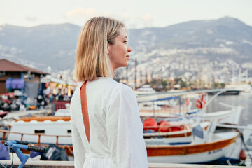 Fototapeta na wymiar Blonde woman in white dress, sea, boats, mountains on the background 