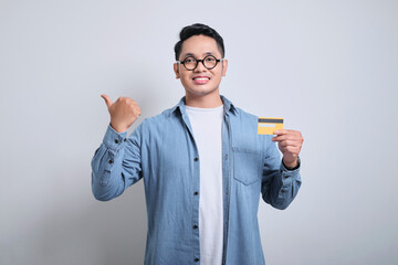 Portrait young asian man wearing denim and glasses smiling happy and his right thumb pointing right side while showing credit card. Payment, debit, credit card concepts.