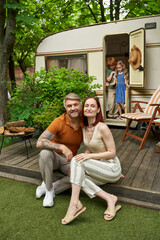 joyful husband and wife sitting and looking at camera near children in trailer home, family leisure