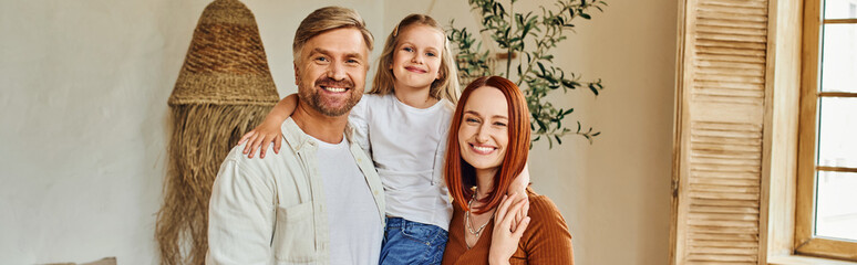 smiling husband and wife holding cute daughter and looking at camera in cozy bedroom, banner