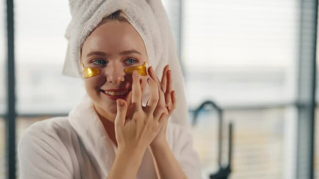 Smiling woman applying gel eye patches. Positive girl in white bathrobe, towel on head making eye skincare beauty procedure in bathroom after shower at home. Spa, wellness, self care bodycare concept.