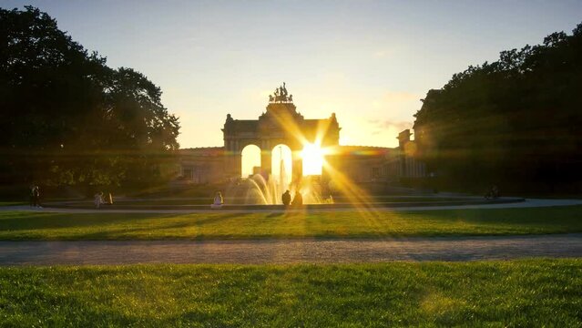 Romantic sunset time lapse of the Cinquantenaire Park with the fountain and the lights turning on as the sun goes down. Lovers and visitors having a picnic in the large park with clouds and many color