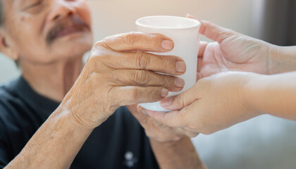 Cropped image hands of caregiver giving senior man with a cup of coffee or tea for breakfast in...