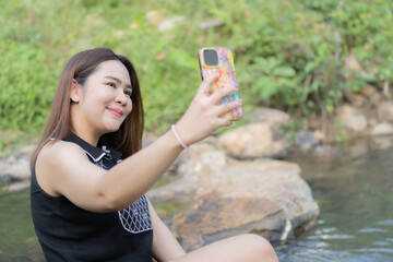 Smiling Asian female taking selfie by smartphone along sunny summer stream.