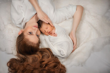 A beautiful girl with long red hair hugs her newborn baby with her arms.
Mom gently touches her newborn baby. Portrait of a newborn and his mother. Newborn baby is sleeping. New Year. Christmas. Son. 