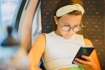 Small beautiful girl in glasses sits on train, using her phone