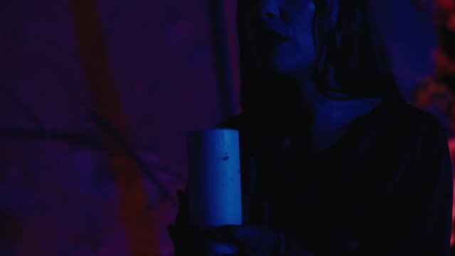 A gorgeous witch woman, with dark makeup and dressed in a black dress, blows out a candle against a colorful background. Slow motion, outdoors during the night.