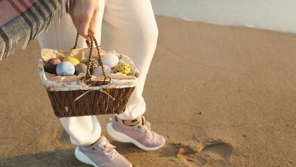 I'm standing on the sand by the sea and holding a wicker brown basket with Painted Easter Eggs. - obrazy, fototapety, plakaty