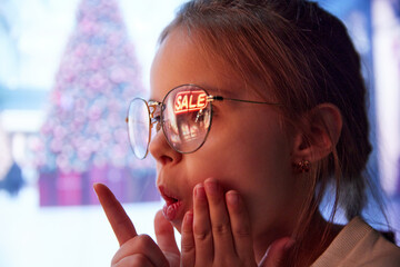 Little girl feeling excited about big sales season. Word sale reflecting on kid's glasses. Winter shopping. Concept of Christmas, childhood, dreams, fantasy, happiness, inspiration