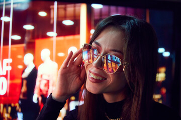 Happy and excited young woman looking on shop windows with neon lights reflection on glasses....