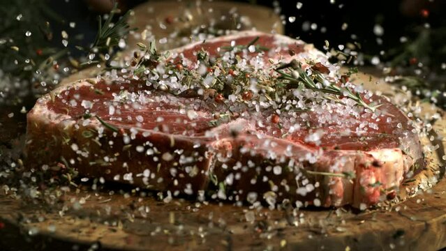 Extreme slow motion macro of bio juicy beef steak on rustic wooden cutting board with mixed herbs spices and salt grains falling on rustic table in kitchen of restaurant before being cooked by chef.