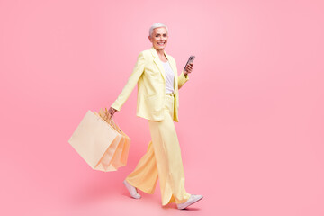 Full size photo of cheerful woman dressed yellow suit go to empty space holding shopping bags...