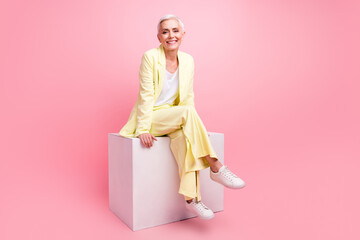 Full size photo of good mood nice cheerful woman dressed yellow suit sit on white cube legs crossed isolated on pink color background