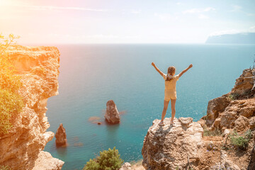 Happy girl stands on a rock high above the sea, wearing a yellow jumpsuit and sporting braided...