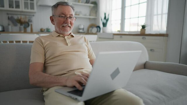 Senior man happy about paying last mortgage payment on laptop using bank credit card. Overjoyed man clenches fists says yes sits on couch in living room. Mortgage repayment, loan repayment concept.