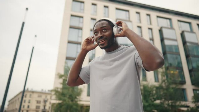 Positive guy listens music on street. African american bearded man in headphones smiling enjoying favourite tracks walking outdoors. Guy in t-shirt in downtown. Melomaniac, music fan lover concept.