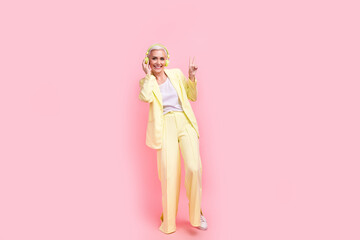 Full body photo of good mood person dressed yellow jacket pants touching headphones showing v-sign isolated on pink color background