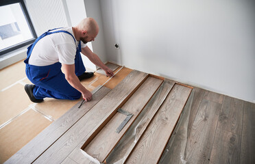 Man in work overalls using tape measure while installing laminate flooring in apartment under...