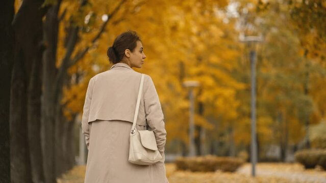 back view of an attractive brunette woman wearing coat walking in park at autumn day with yellow dry leaves on trees. happy beautiful woman in autumn outdoors. Slow-motion 4k footage