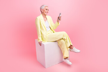 Full body photo of intelligent person dressed yellow jacket pants sit on podium look at smartphone isolated on pink color background