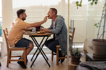 Happy couple of gay men have breakfast together for Valentine's Day while tenderly feeding each other - obrazy, fototapety, plakaty