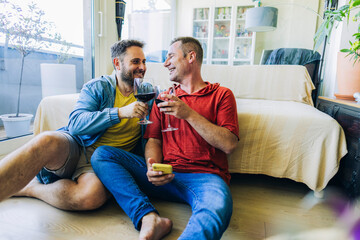 gay couple sitting on the floor toasting with glasses of wine while checking their social networks