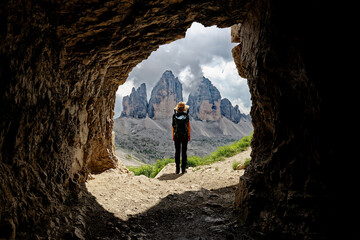 A female person admiring the view of Tre Cime di Lavaredo mountain in the Dolomites, Italy. Famous places for hiking. View from inside of a cave.  - obrazy, fototapety, plakaty