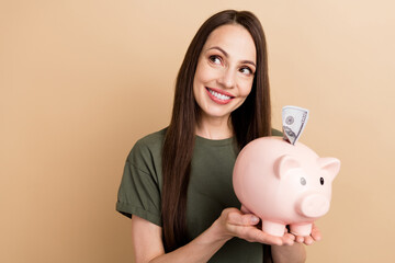 Portrait of dreamy mature woman hold piggy bank looking curious novelty puts banknote inside box isolated on beige color background