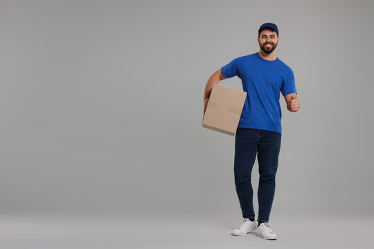 Happy young courier with parcel showing thumb up on grey background, space for text