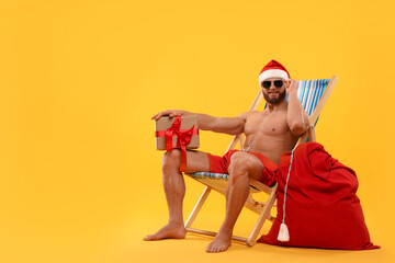 Muscular young man in Santa hat with deck chair, bag, sunglasses and Christmas gift box on orange...