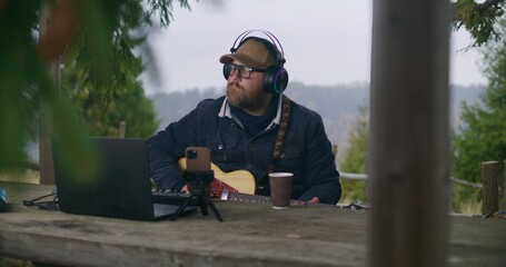 Caucasian man in headphones plays guitar sitting in wooden gazebo in the forest. Male musician...