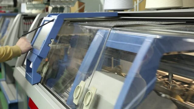 The operator sets up a modern digital knitting machine. A row of industrial textil flat knitting machines in a knitwear factory. Manufacture of knitwear, modern computerized flat Knitting Machine