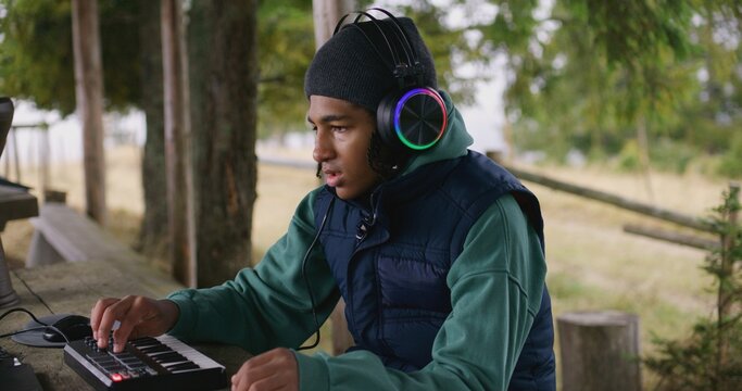 African American teenager wearing headphones plays on MIDI controller outdoors. Young musician, composer creates music, mixes songs, uses laptop for recording while sitting in gazebo in the forest.