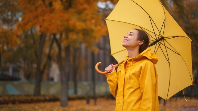 girl take off yellow raincoat hoodie to enjoy falling rain, standing with umbrella in city park looking up to sky enjoying rain drops to her face smiling, feel fresh, season climate change