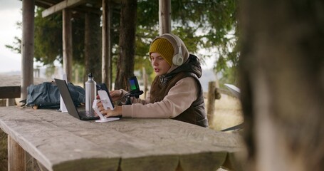 Teenager in headphones uses laptop and phone on tripod to create content for viewers during holidays in mountain forest. Young streamer sits in wooden gazebo and talks into professional microphone.