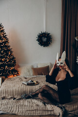 Young playful woman sitting on floor in decollete dress and bunny mask against background of Christmas tree.
