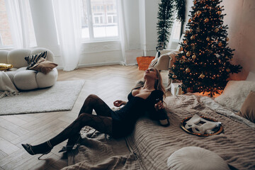 Young woman sitting on floor and relaxing in decollete dress with wine bottle against Christmas tree.