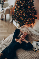Young pensive blonde woman lying down on bed in decollete dress near Christmas tree.